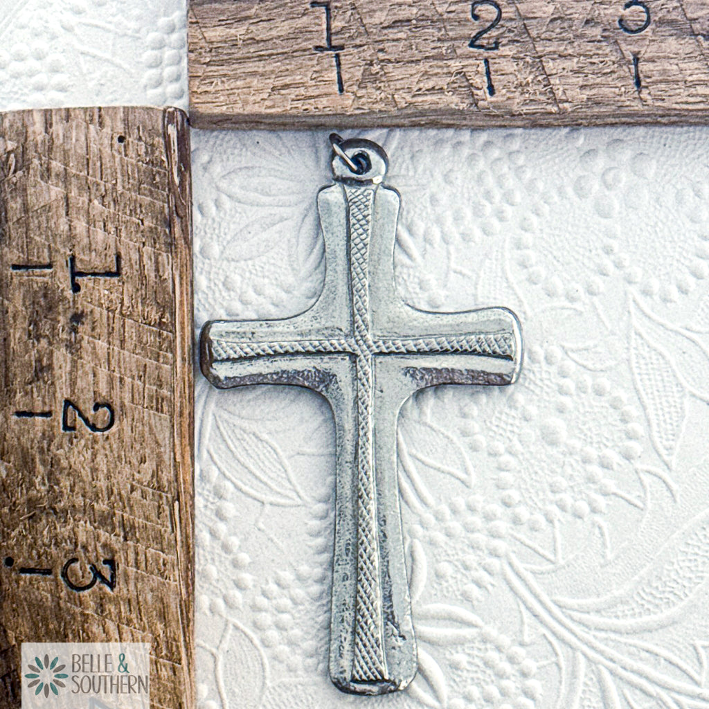 Silver cross pendant on a textured white surface with a wooden ruler for scale.