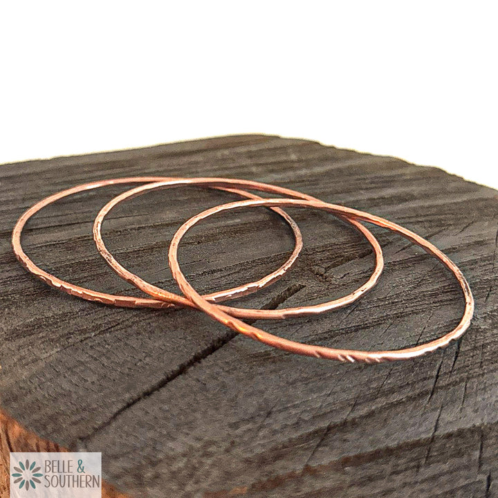 Set of three handforged recycled copper bangles, one scalloped, one lined and one hammered, resting on dark charred wood