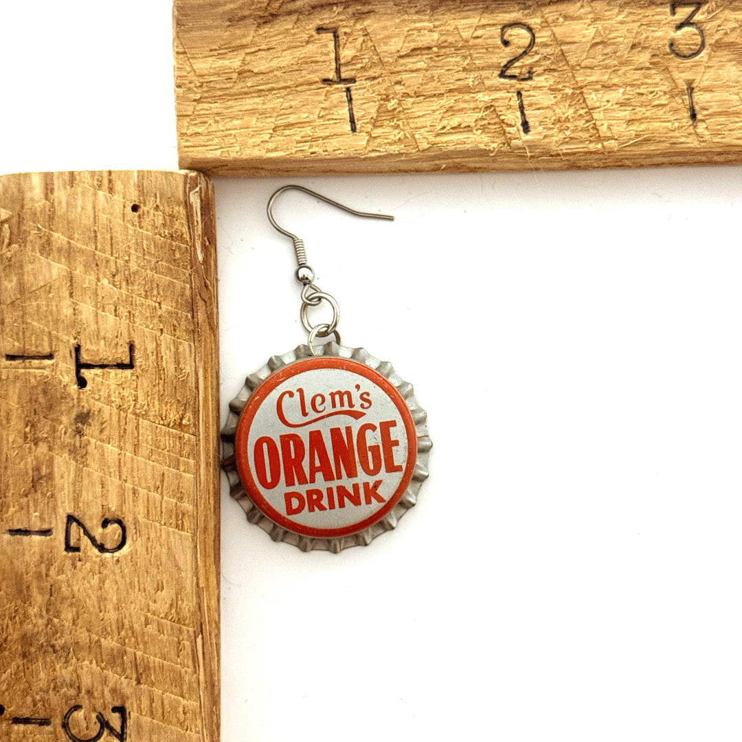 Silver and Orange Clem's Orange Drink Bottle Cap on Steel Ear Wire Earrings next to ruler