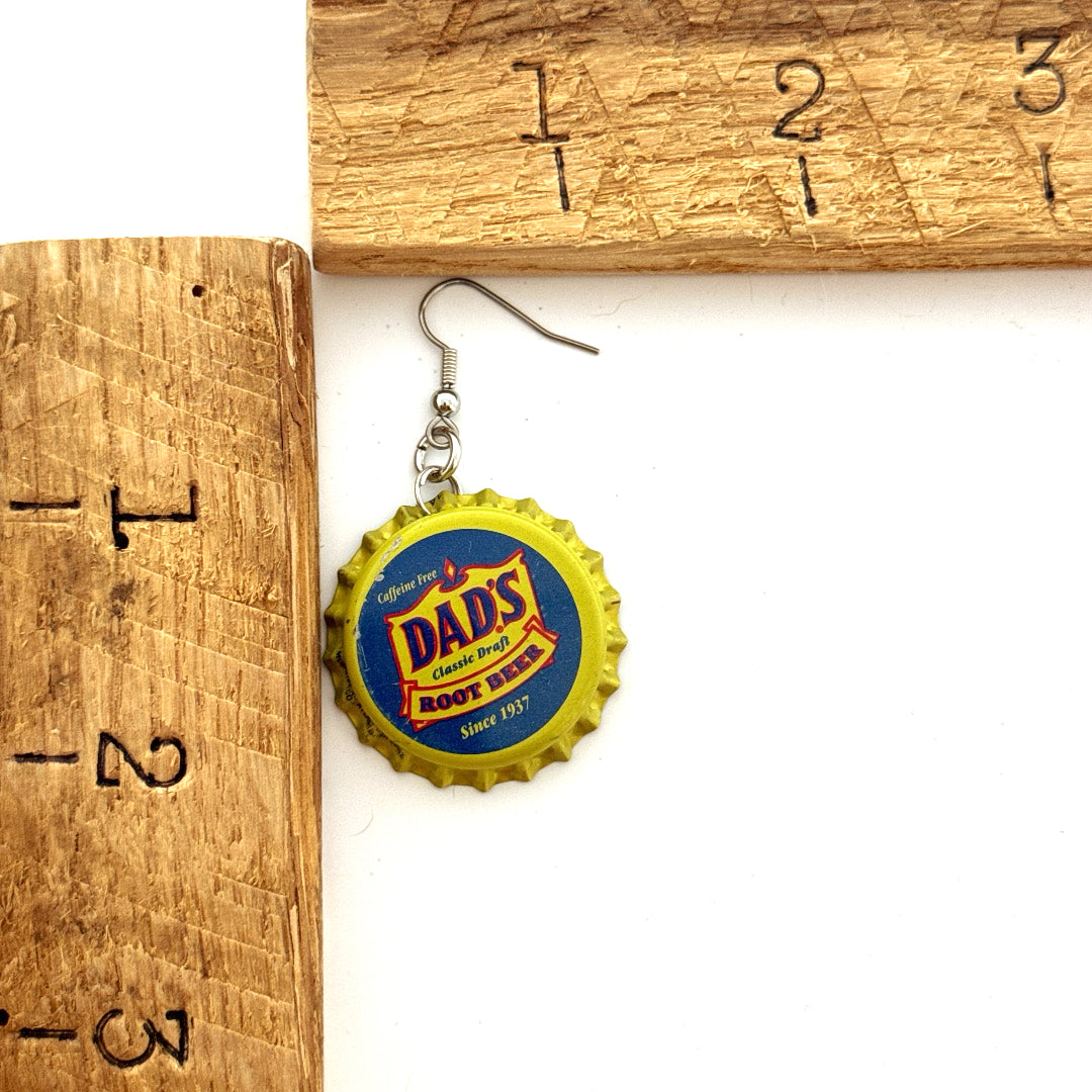 Yellow and Blue Dad's Root Beer Bottle Cap on Stainless Steel Ear Wire Earrings next to ruler