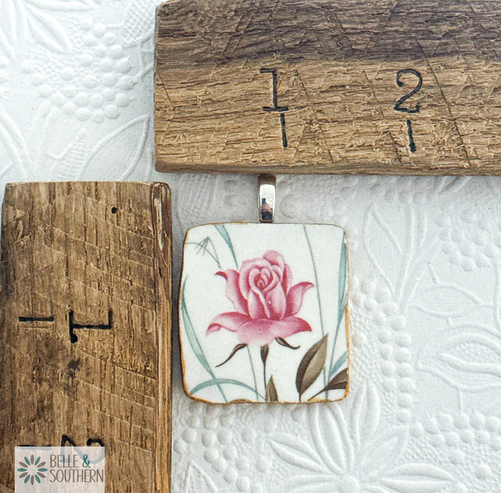 Decorative ceramic tile with pink rose design on a textured white surface with wooden ruler for scale.