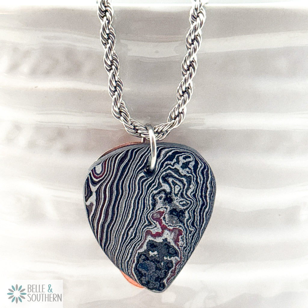 Fordite Guitar Pick shaped  with left side features tight, parallel waves of white, charcoal, and slate-blue, while the right side shifts into organic, molten swirls resting on copper backplate with Stainless Steel rope chain
