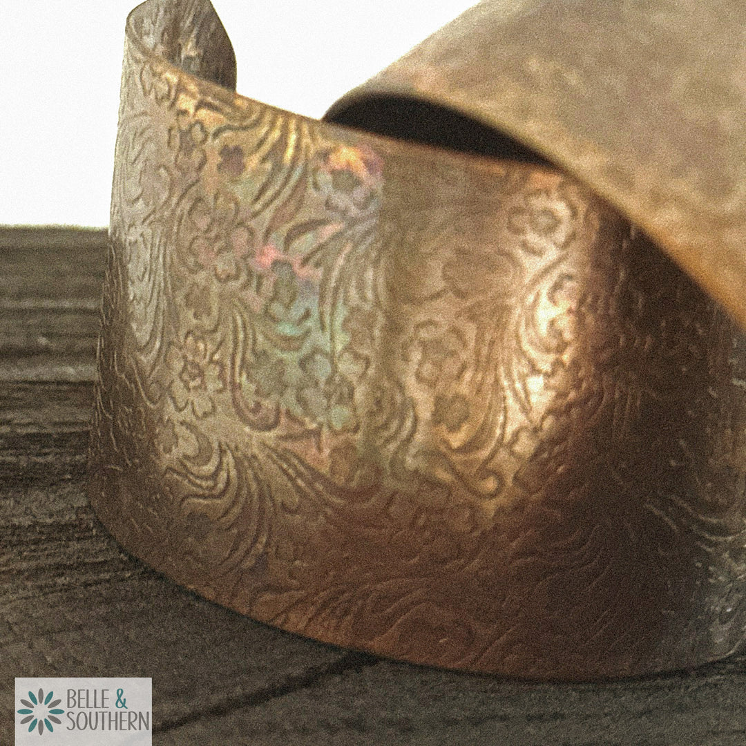 Close-up of a handforged copper cuff bracelet with small blossoms and feathered scrollwork pressed into the metal, warm gold with pink and green iridescent flashes, resting on weathered grey wood