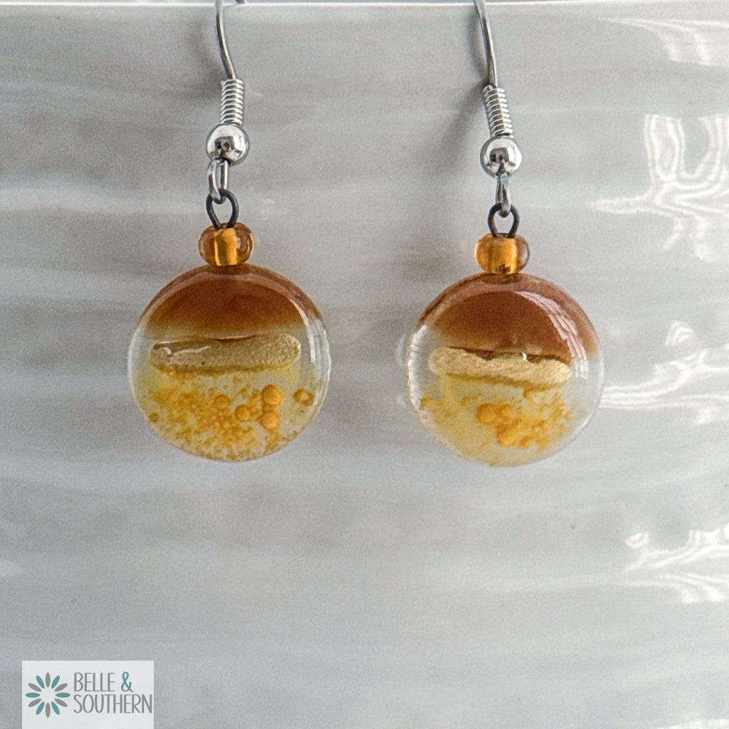 Amber and Gold circular fusion glass earrings with a stainless steel ear wire  on a white vase, branded 'Belle & Southern'.