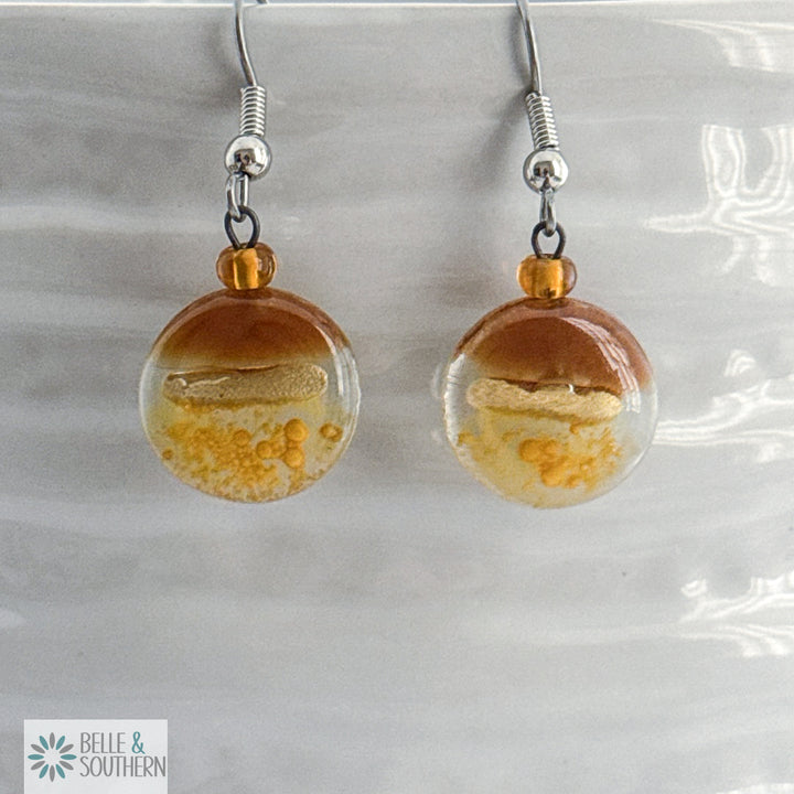 Amber and Gold circular fusion glass earrings with a stainless steel ear wire  on a white vase, branded 'Belle & Southern'.