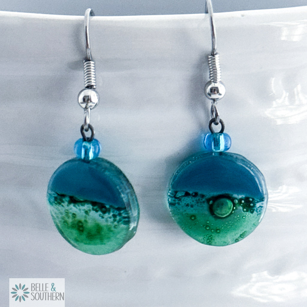 Pair of blue and green fusion glass earrings on a white vase with 'Belle & Southern' branding.