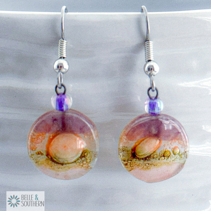 Lavender, Gold and Pink circular fusion glass earrings with a stainless steel ear wire  on a white vase, branded 'Belle & Southern'.