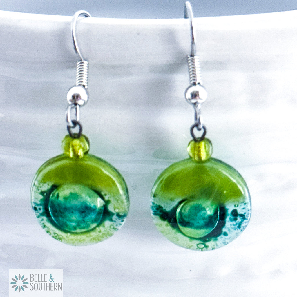 Green circular fusion glass earrings with a stainless steel ear wire  on a white vase, branded 'Belle & Southern'.