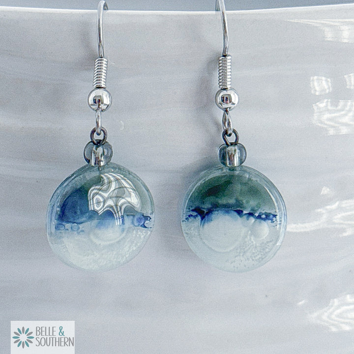 Grey, blue and white circular fusion glass earrings with a stainless steel ear wire  on a white vase, branded 'Belle & Southern'.