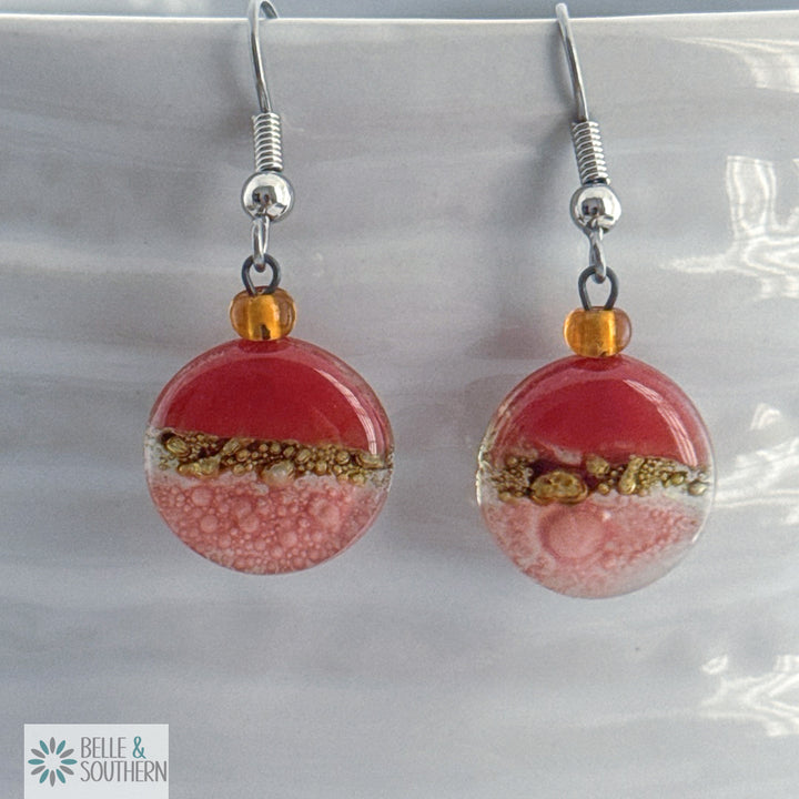 Coral, gold and pink circular fusion glass earrings with a stainless steel ear wire  on a white vase, branded 'Belle & Southern'.