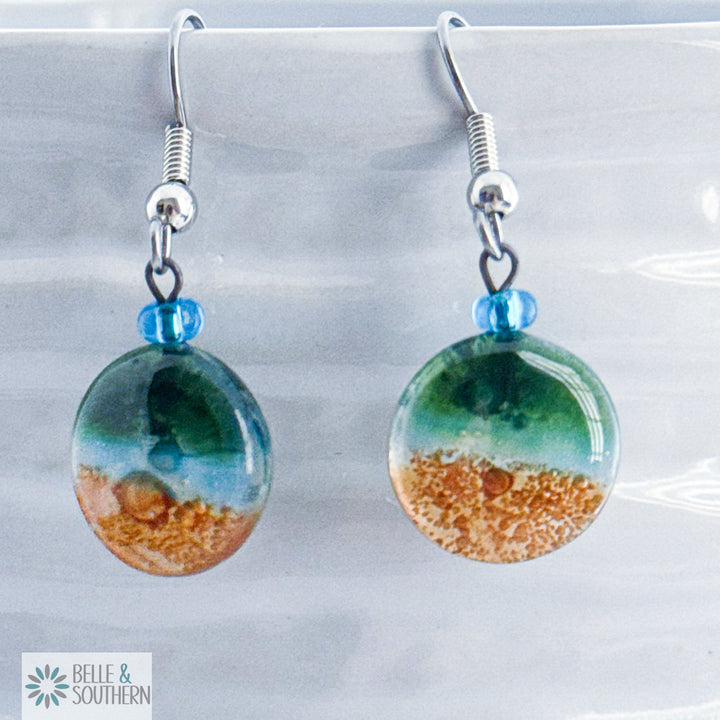 Green, light blue and gold circular fusion glass earrings with a stainless steel ear wire  on a white vase, branded 'Belle & Southern'.