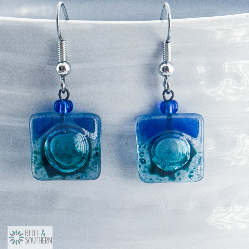 Dark blue and light blue fused glass square earrings on stainless steel ear wires on a porcelain vase
