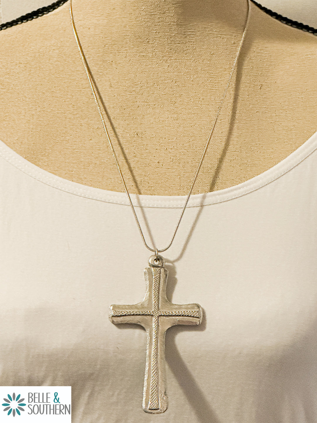 A silver-colored cross pendant displayed on a mannequin wearing a white top.