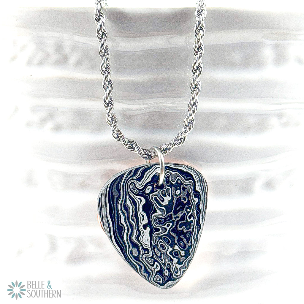 Grey, black, Silver , white and subtle blue guitar pick shape resting on a copper backplate.  On a stainless steel rope chain hanging on a white vase.