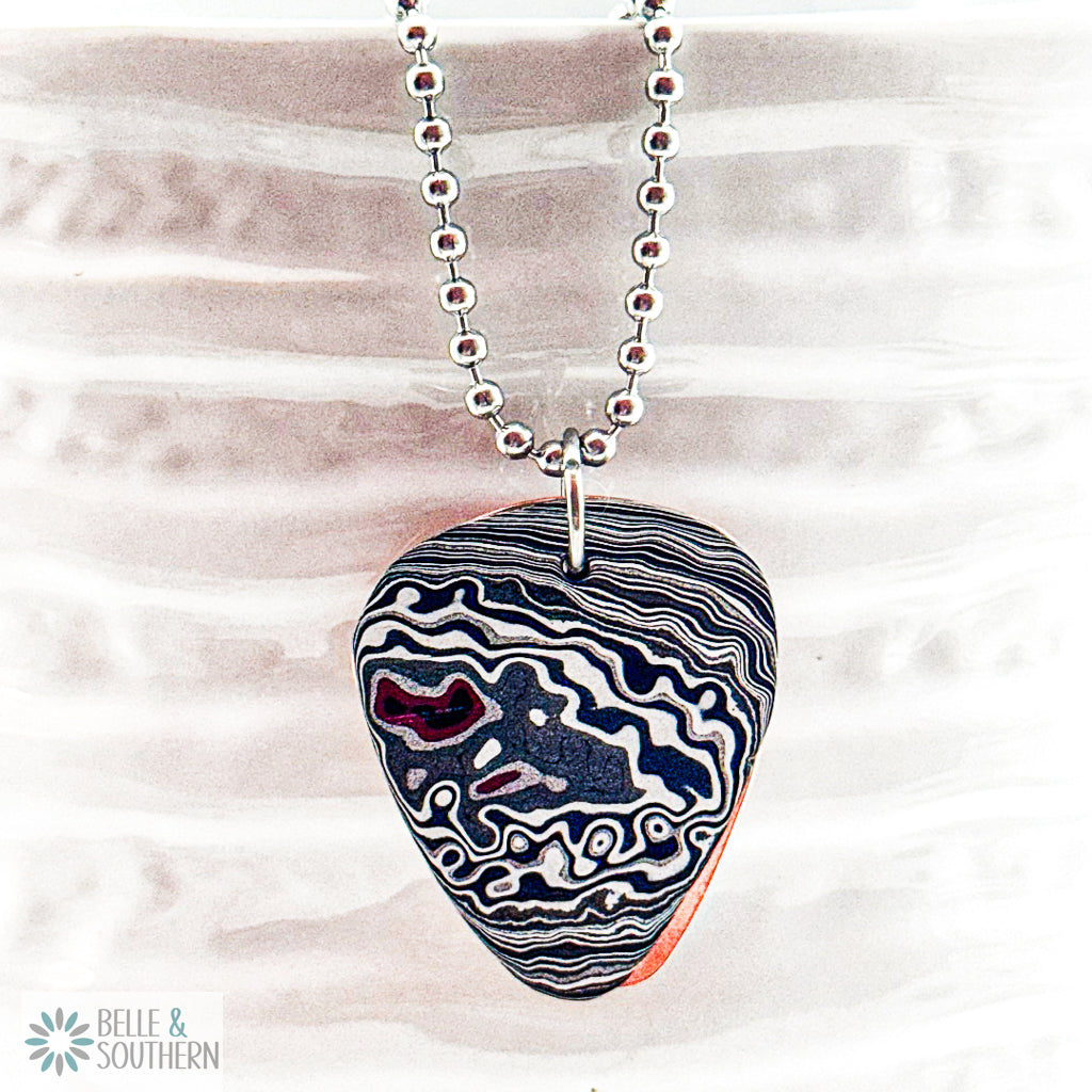Fordite Guitar Pick Shaped resting on copper backplate on stainless steel ball chain hanging on white vase