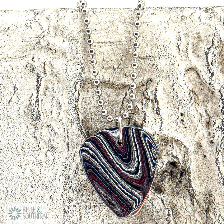 Fordite Guitar Pick Necklace on a bead chain up against a stone surface
