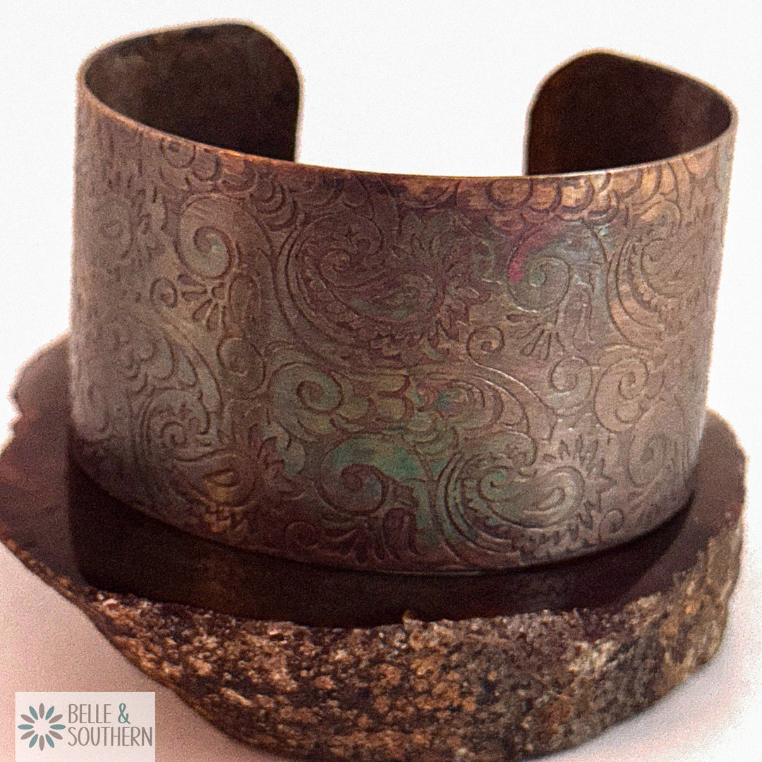Wide handforged copper cuff bracelet with a paisley pattern pressed into the metal, hand-darkened to an iridescent teal and lavender finish, standing upright on a stone slab