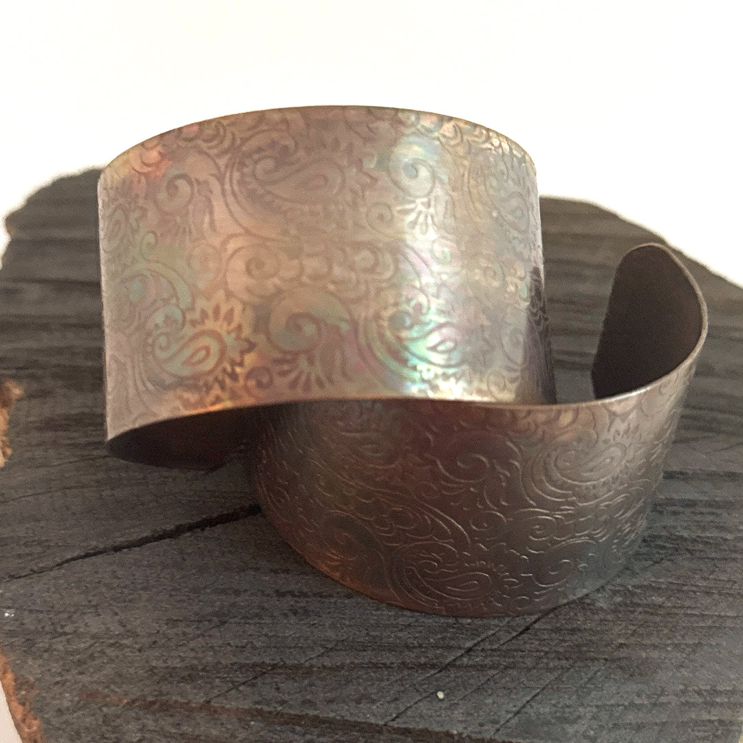 Iridescent recycled copper paisley cuff bracelet resting on dark wood, showing rose, green and teal flashes across the pressed teardrop pattern