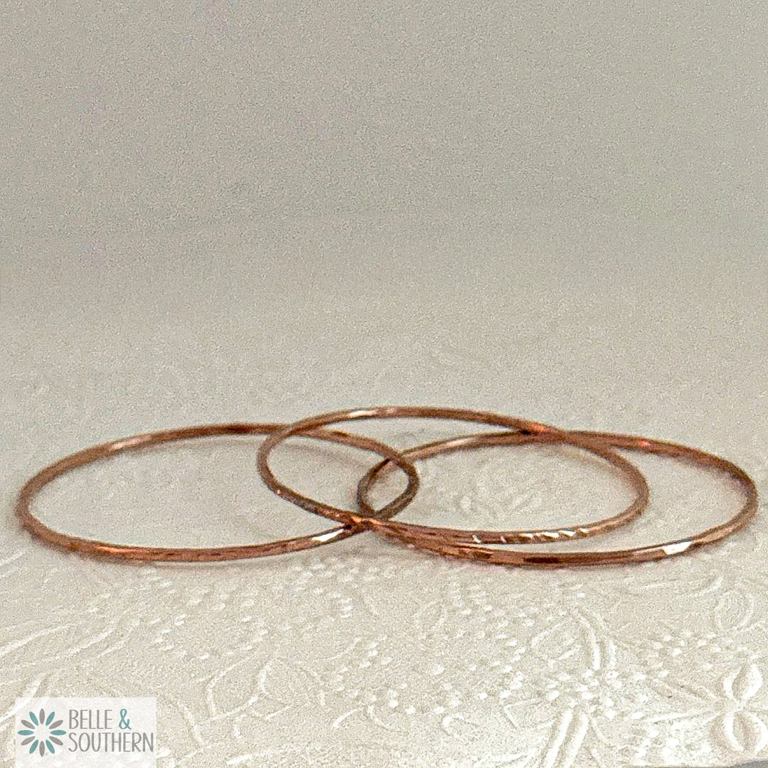 Three handforged recycled copper bangles laid in a row on a cream embossed floral background, one scalloped, one lined and one hammered