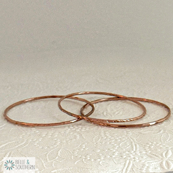 Three handforged recycled copper bangles laid in a row on a cream embossed floral background, one scalloped, one lined and one hammered