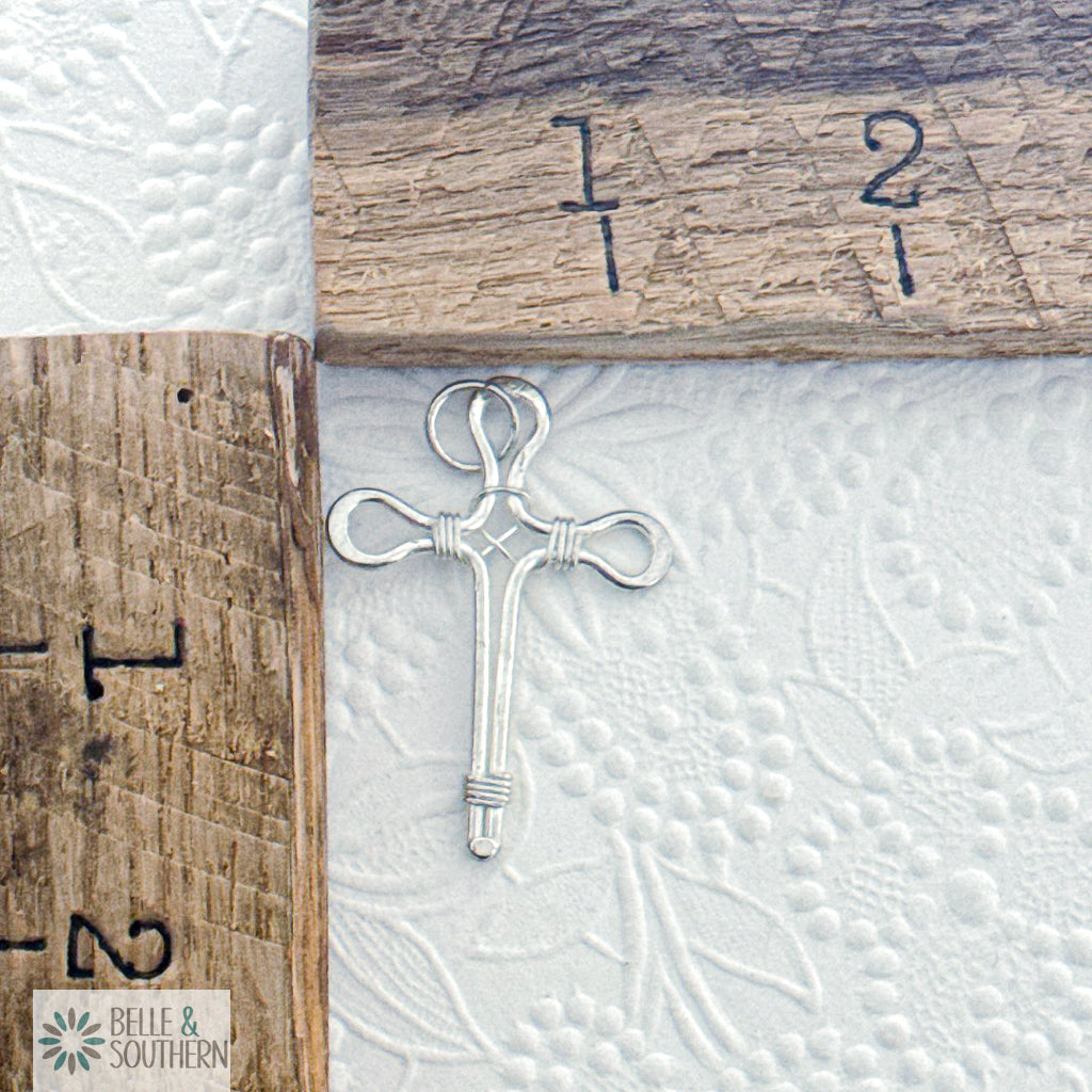 Silver cross pendant on textured white by wooden rulers for scale with 'Belle & Southern' branding.