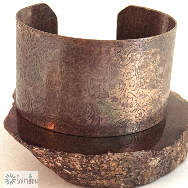 Wide recycled copper cuff bracelet with a floral pattern pressed into the metal, hand-darkened to an iridescent lavender and rose finish, standing open on a rough stone slab
